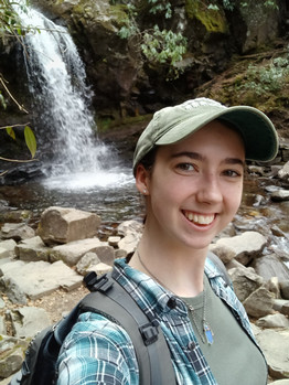 Brianne Brummett takes a selfie in front of a waterfall in a green cap and ponytail.