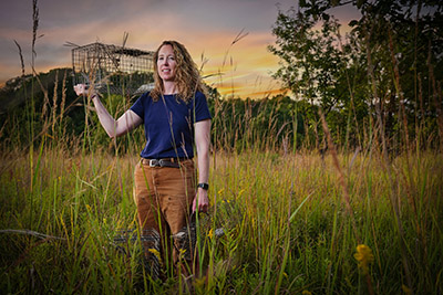 Dr. Elizabeth Flaherty stands in a prairie while holding a trap on her shoulder.