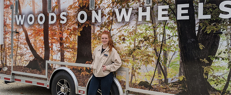 Sara Miller, alumna, standing in front of travel exhibit Woods on Wheels.