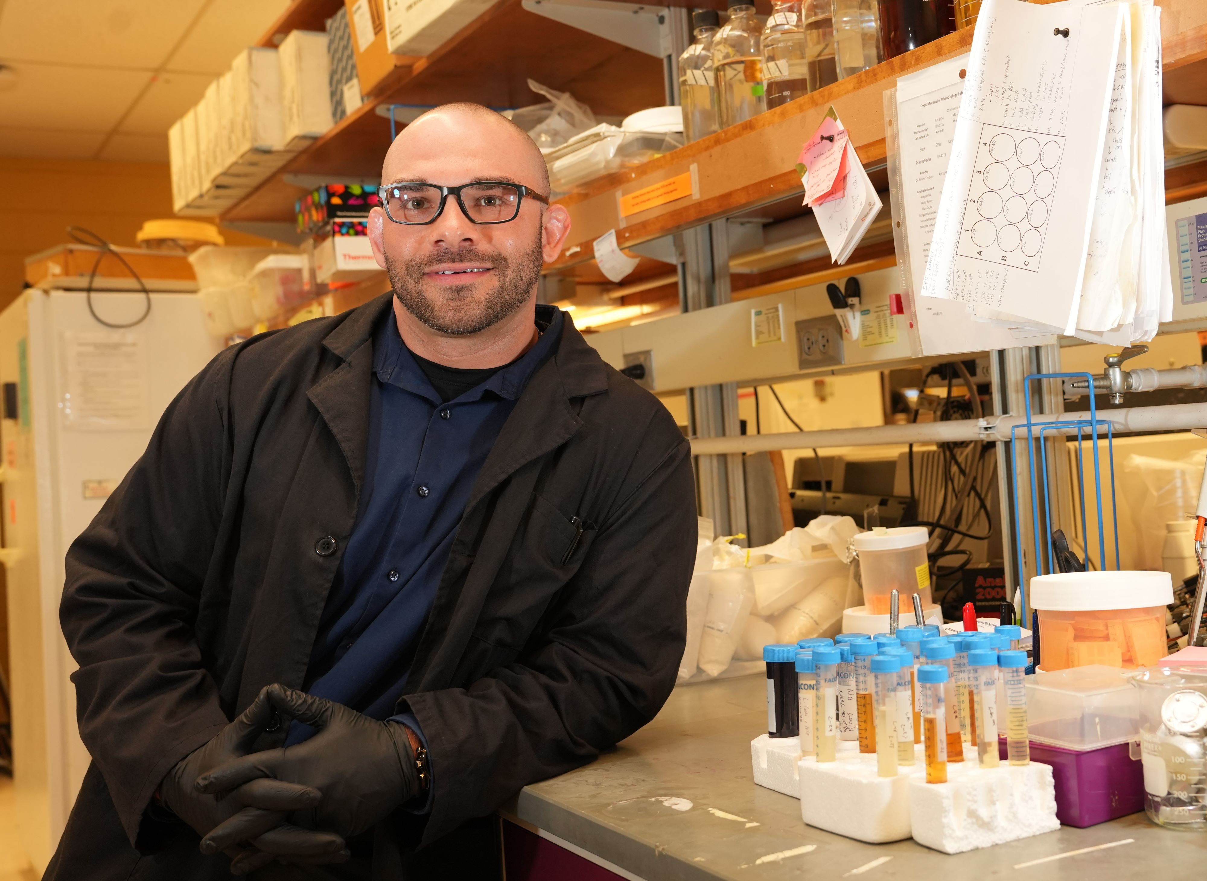 Man wearing a lab coat and gloves while leaning on a lab benchtop next to test tubes