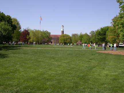 Large open lawn on a university campus with scattered people, trees, and a building with a clock tower in the background.