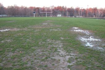 Heavily worn athletic field with patchy grass, exposed soil, and muddy areas in high‑traffic zones near goalposts.