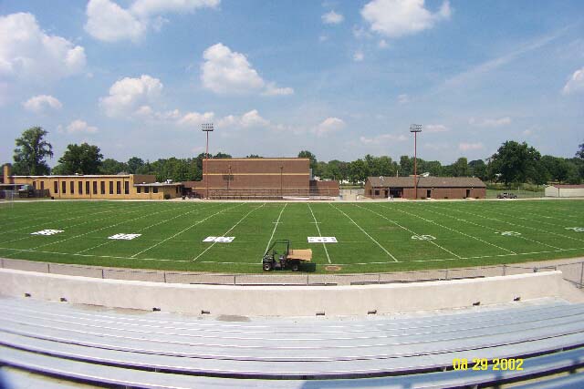 Overseeded bermudagrass football field with uniform green turf, yard line markings, stadium seating in the foreground, and school buildings in the background.