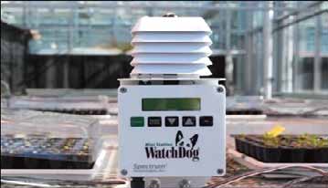 WatchDog weather station with a radiation shield and digital display positioned among greenhouse benches. The portable monitoring device is used to measure environmental conditions and calculate daily light integral (DLI) for greenhouse crops.