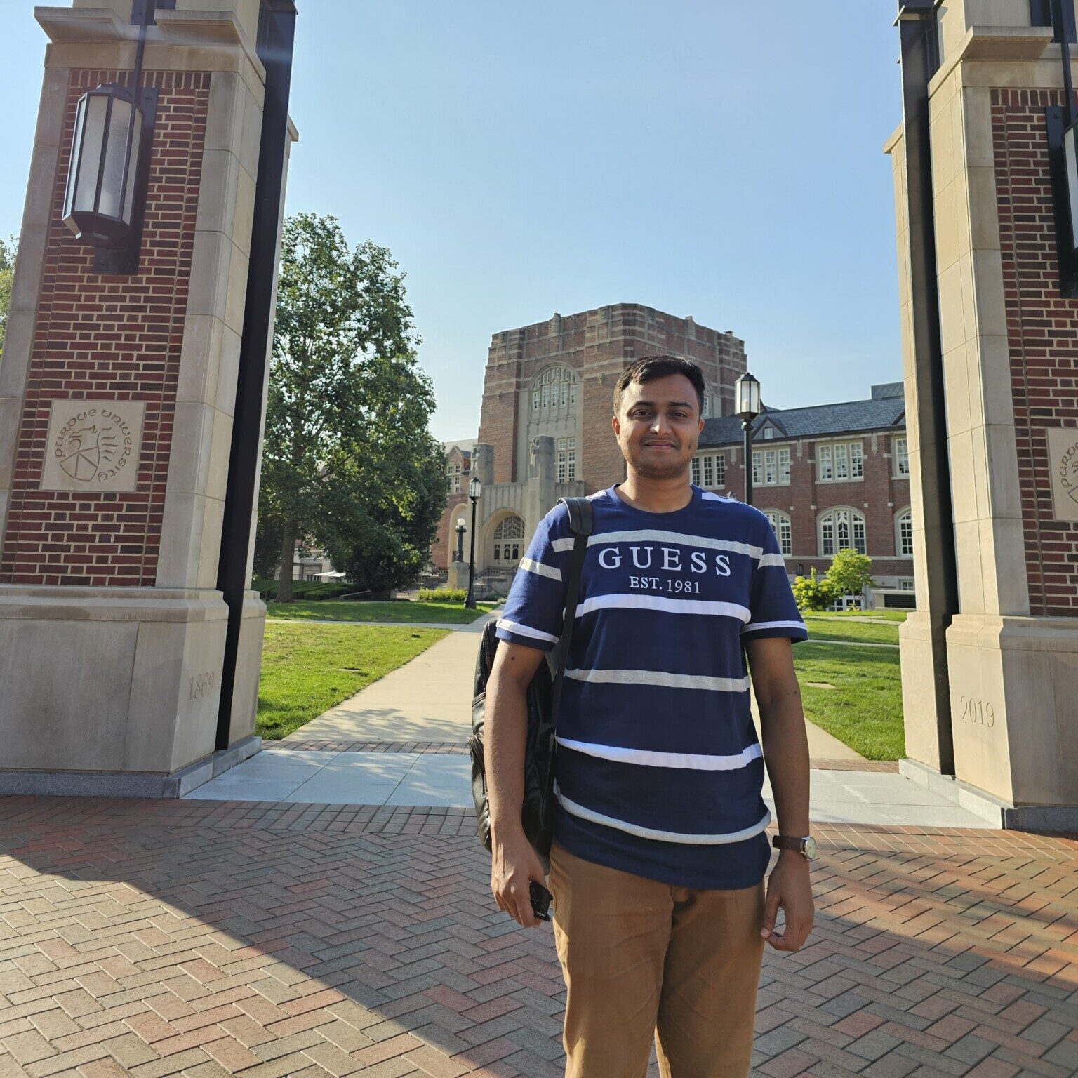 Graduate student standing on Purdue University’s campus between two brick entrance pillars.