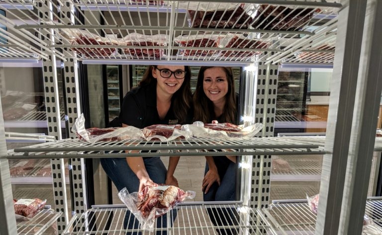 women behind a refrigerated compartment showing meat products  