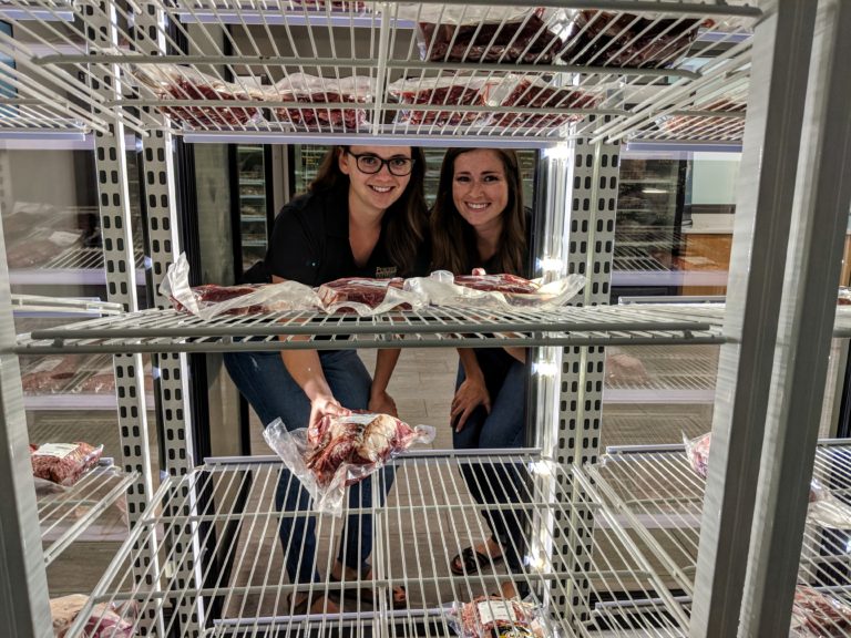 women behind a refrigerated compartment showing meat products  