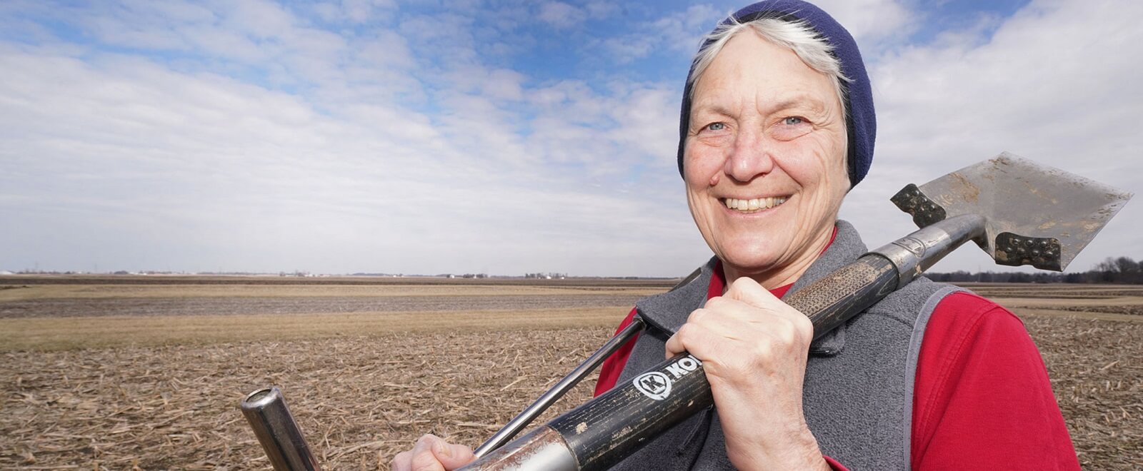 Kladivko smiling as she hold a farming tools resting on her shoulder (outdoors on a cornfield). 