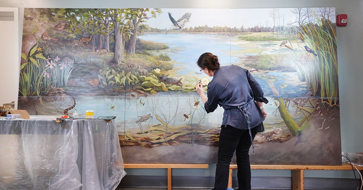 Woman painting a wetland ecosystem that includes birds, reptiles, bugs, other animals and flowers