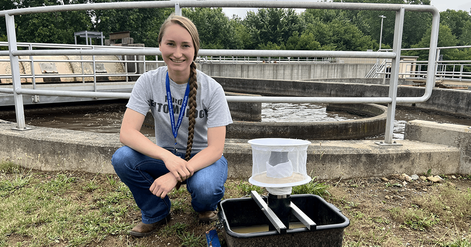 Student Lexi LaMar poses and squats next to a "gravid trap" with a concrete pool of water from a wastewater treatment plant behind her