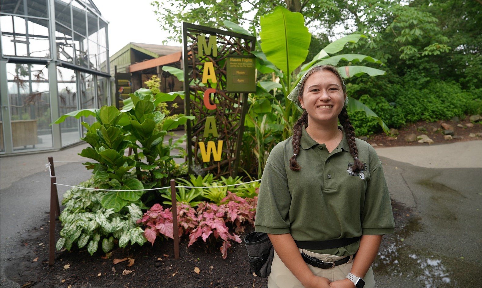 Student Emily Carlisle stands in a green polo next to a sign with letters that spell "macaw" in front of a macaw enclosure