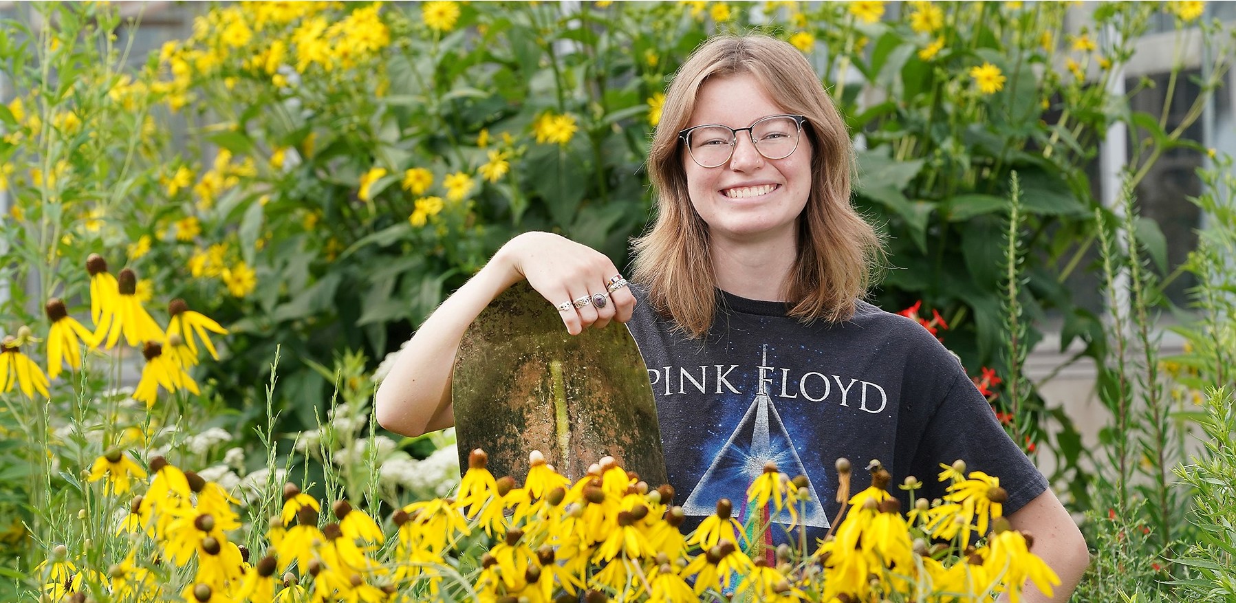 Mackenzie Sandusky poses in front of tall yellow flowers leaning on a shovel