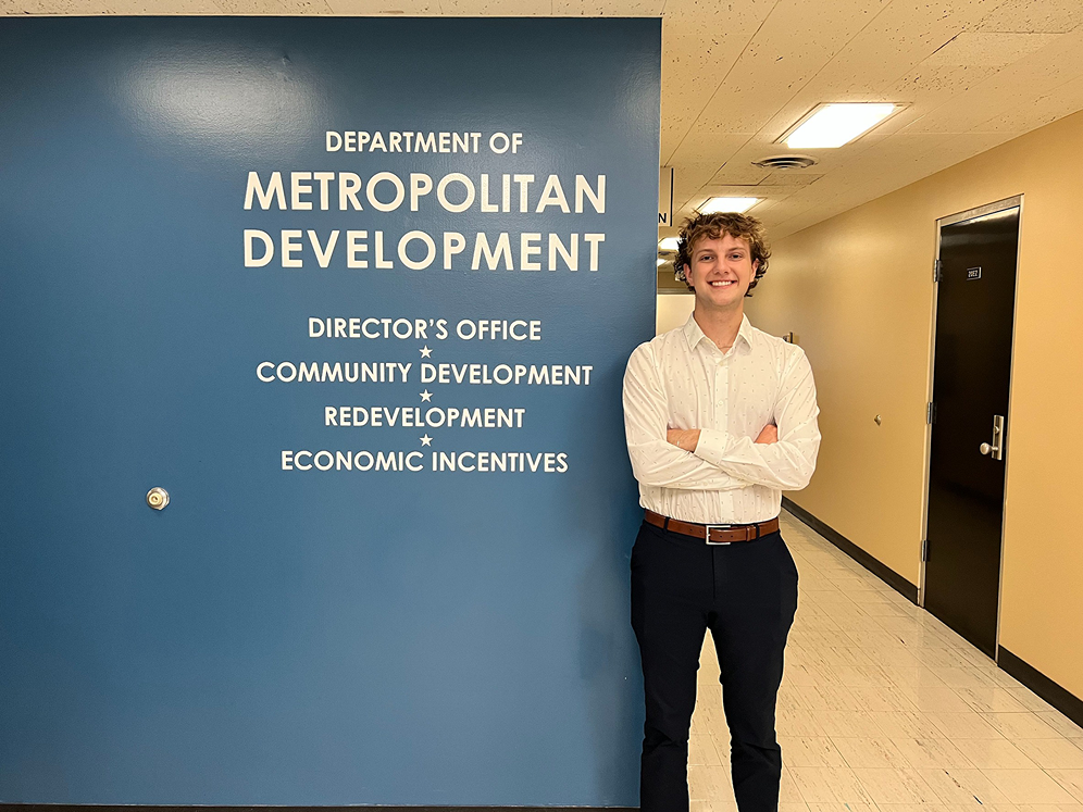 Nicholas Neuman poses in front of a blue wall that says "Department of Metropolitan Development"