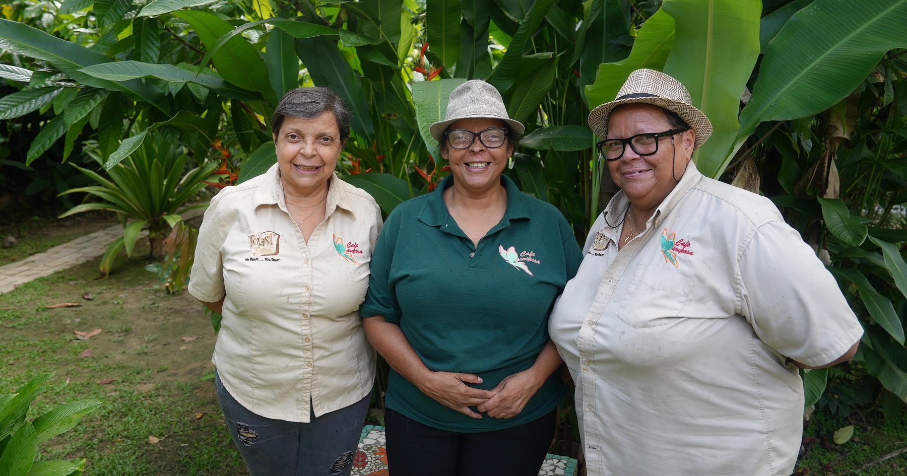 Three Café Mariposa sisters smile and stand in front of green large-leafed tropical plants. They wear tan collared shirts that have a butterfly logo and the words Café Mariposa above their left pocket. Marcia Guerrero is one of the sisters in the picture.