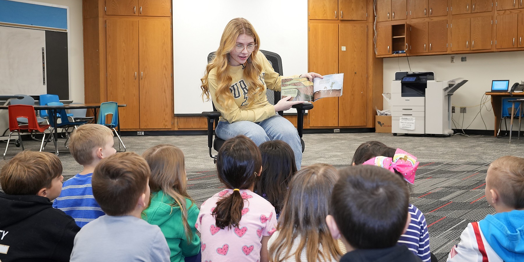 Student Kourtney Otte sits in a chair reading animatedly to a class of kindergarten students