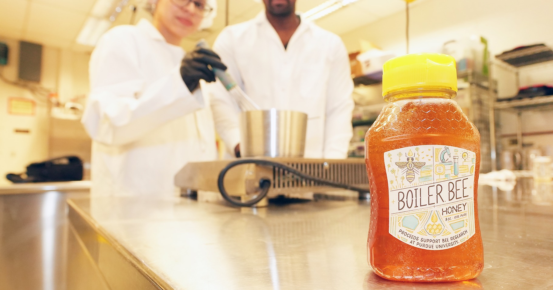 A bottle of Boiler Bee Honey sits on the edge of chrome table in Skidmore lab with two students cooking in labcoats and hairnets in the background.