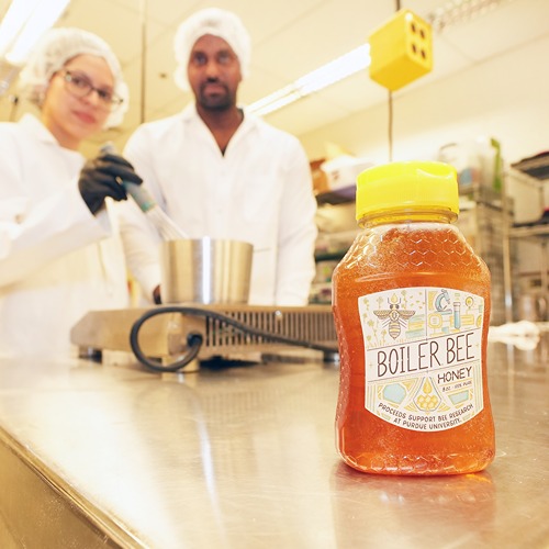 A bottle of Boiler Bee Honey sits on the edge of chrome table in Skidmore lab with two students cooking in labcoats and hairnets in the background.