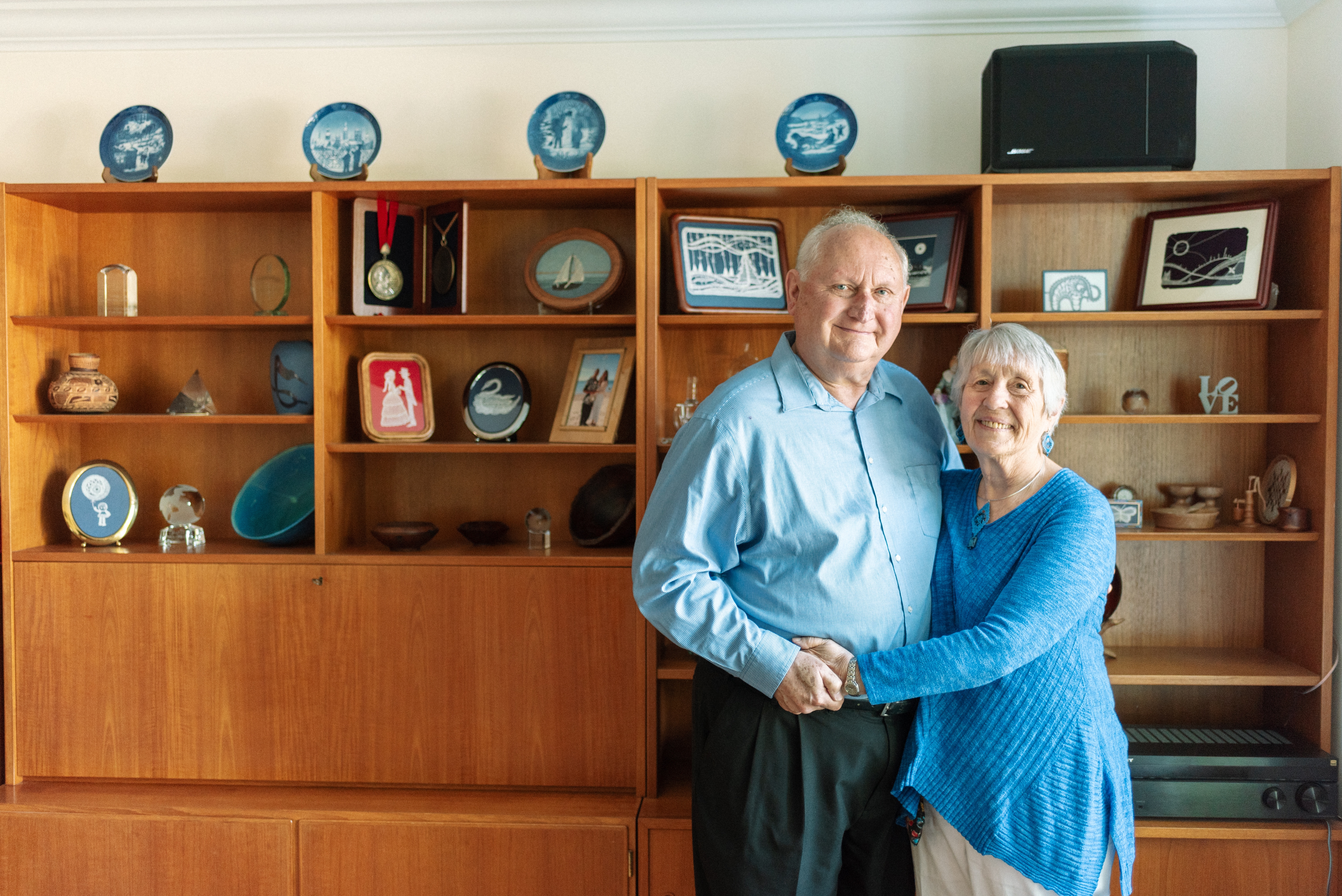 Bob and Karen stand side-by-side while embracing in front of a wooden shelving unit