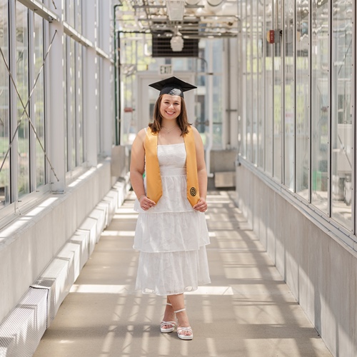 Senior Kayla Grennes stands in cap and sash in Purdue Greenhouse 