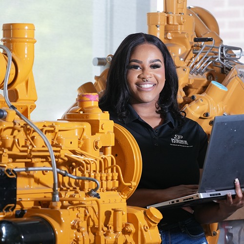Student stands between yellow machinery holding laptop while smiling at the camera