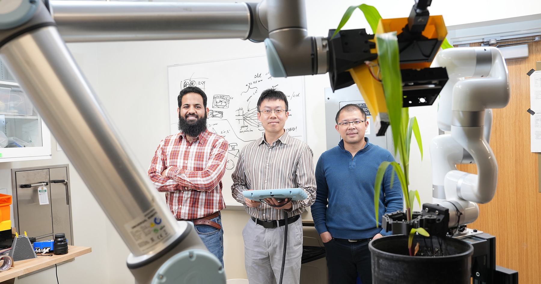 a robotic arm reaches out to touch a corn plant leaf. you can see the Sheeraz Athar, Jian Jin, and Yu She in the where the arm bends