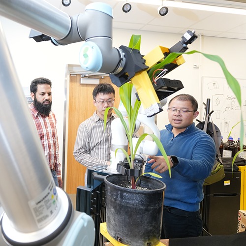 a robotic arm reaches out to touch a corn plant leaf. you can see the Sheeraz Athar, Jian Jin, and Yu She in the where the arm bends