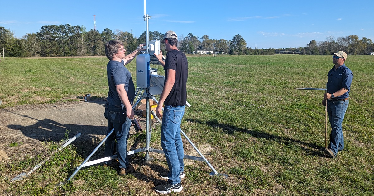 two people tighten a box-shaped device to the SPRING tripod in an open field. A third is coming towards them to help