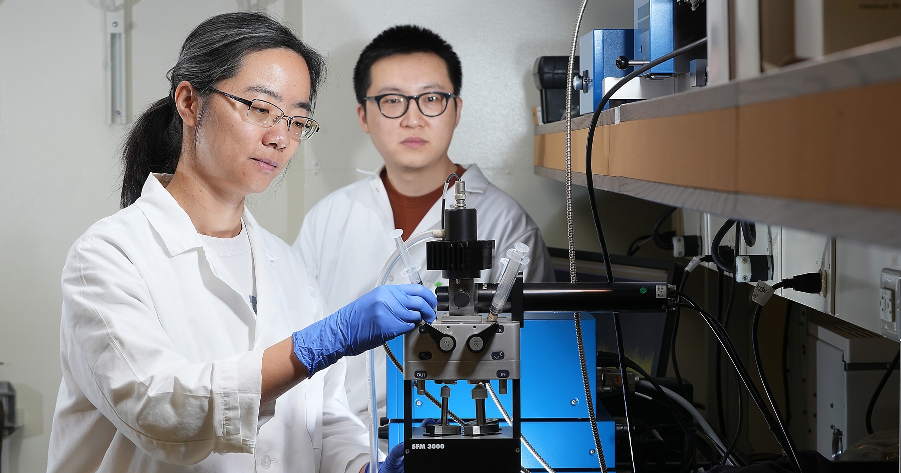 Xing Liu and another lab member stand beside a stopped-flow machine in lab coats