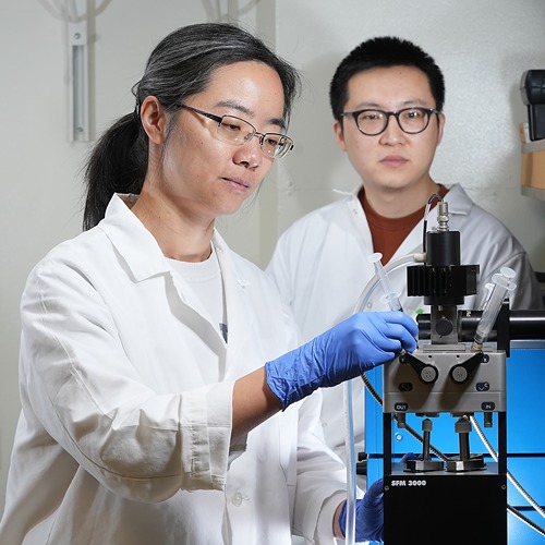 Xing Liu and another lab member stand beside a stopped-flow machine in lab coats