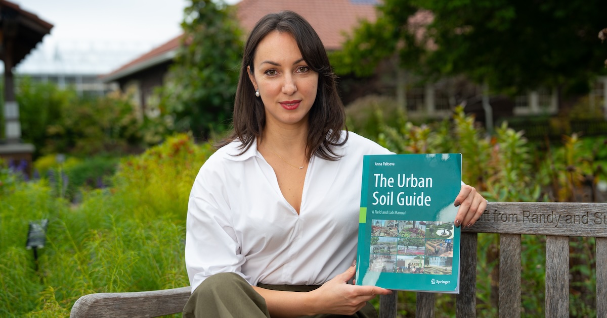 Anna Paltseva, clinical assistant professor of urban science in the agronomy department, sits outside of a greenhouse, holding her Urban Soils Guide.
