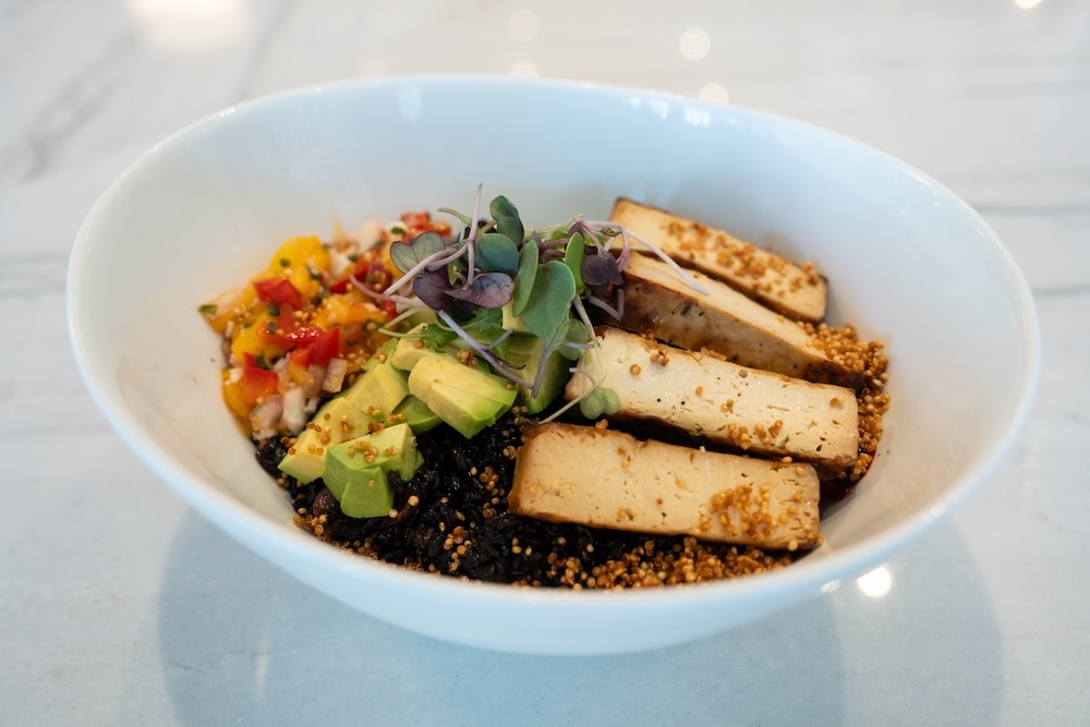 The Forbidden Summer dish - black rice, pan-seared blackened tofu, red onion, avocado, broccoli, mango, cauliflower, toasted quinoa, red pepper and mango chili reduction - plated and sitting on a white marble table.