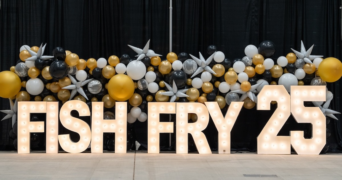 Gold light-up block letters that say, “Fish Fry 2025” in front of a black backdrop and gold, silver and white balloons.