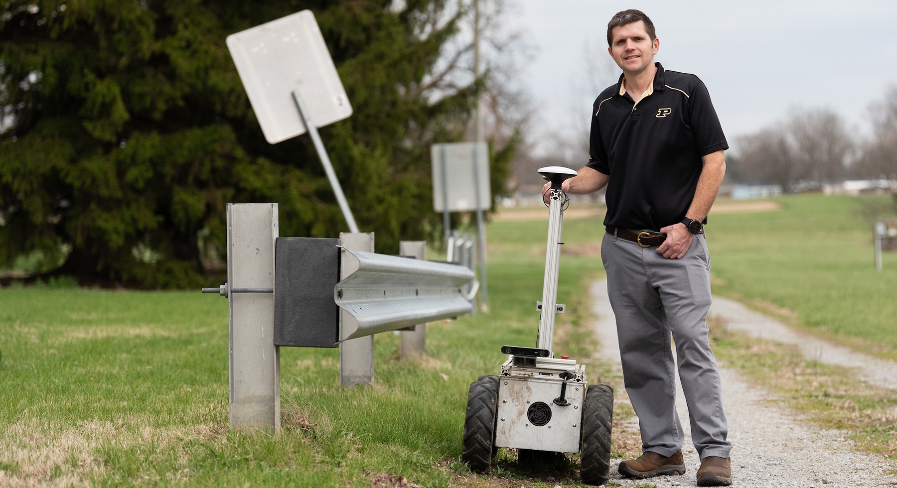 John Evans stands on a gravel road next to an autonomous mower and guard rail