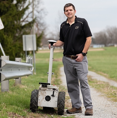 John Evans stands on a gravel road next to an autonomous mower and guard rail