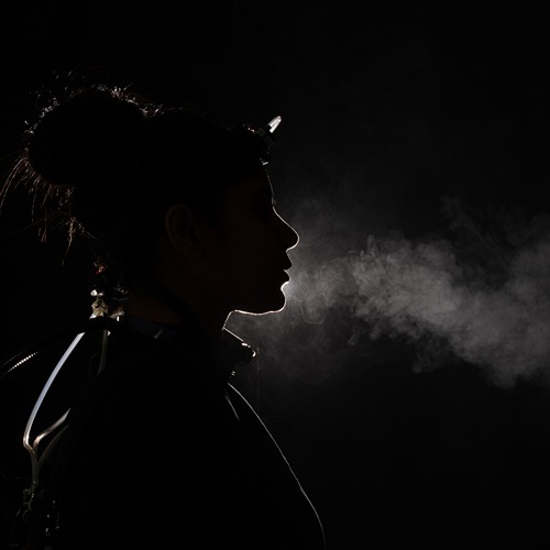 a woman wearing wires and carbon dioxide sensors is lit from behind, her breath making a trail of white clouds in front of her mouth