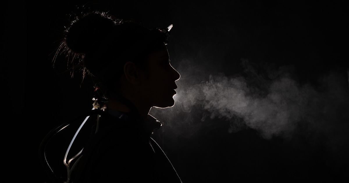 a woman wearing wires and carbon dioxide sensors is lit from behind, her breath making a trail of white clouds in front of her mouth
