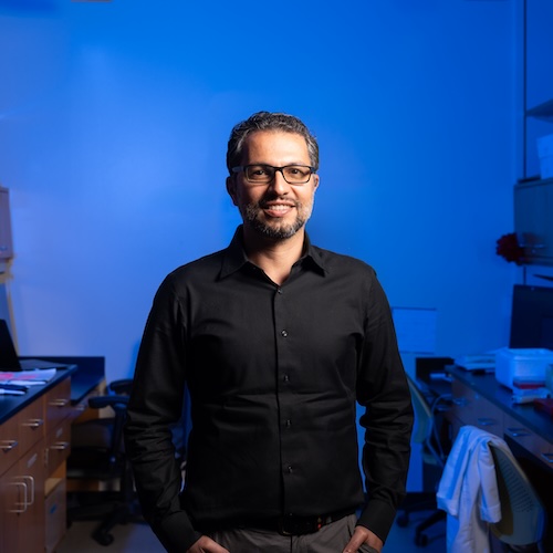 Majid Kazemian in his lab, standing in front of a desk, smiling.