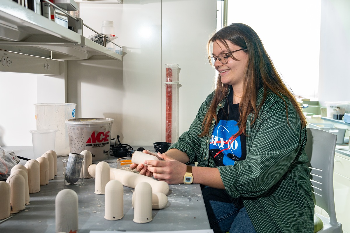 a student in a space shirt smiles and sits at lab bench full of ceramic mycoponic tubes, a small one in her hand