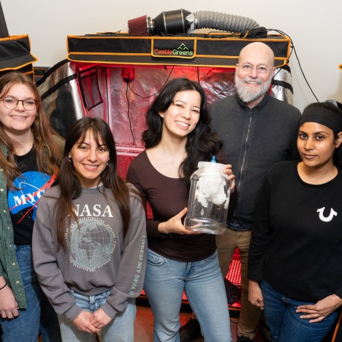 4 students stand in front of marshall porterfield, all in different space and NASA shirts, one carrying a plastic jug with a white fungus growing over a mycoponics tube inside. Grow tents line the background on each side.