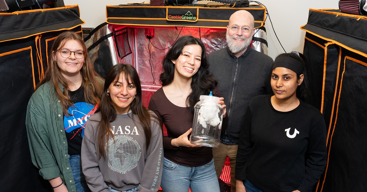 4 students stand in front of marshall porterfield, all in different space and NASA shirts, one carrying a plastic jug with a white fungus growing over a mycoponics tube inside. Grow tents line the background on each side.