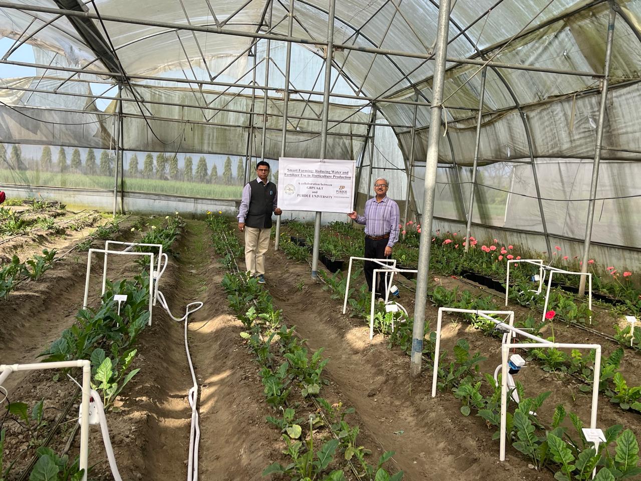 Nemali at the research site in GBPUAT, an outside space with plants and soil, and holding a sign that notes the Purdue collaboration.
