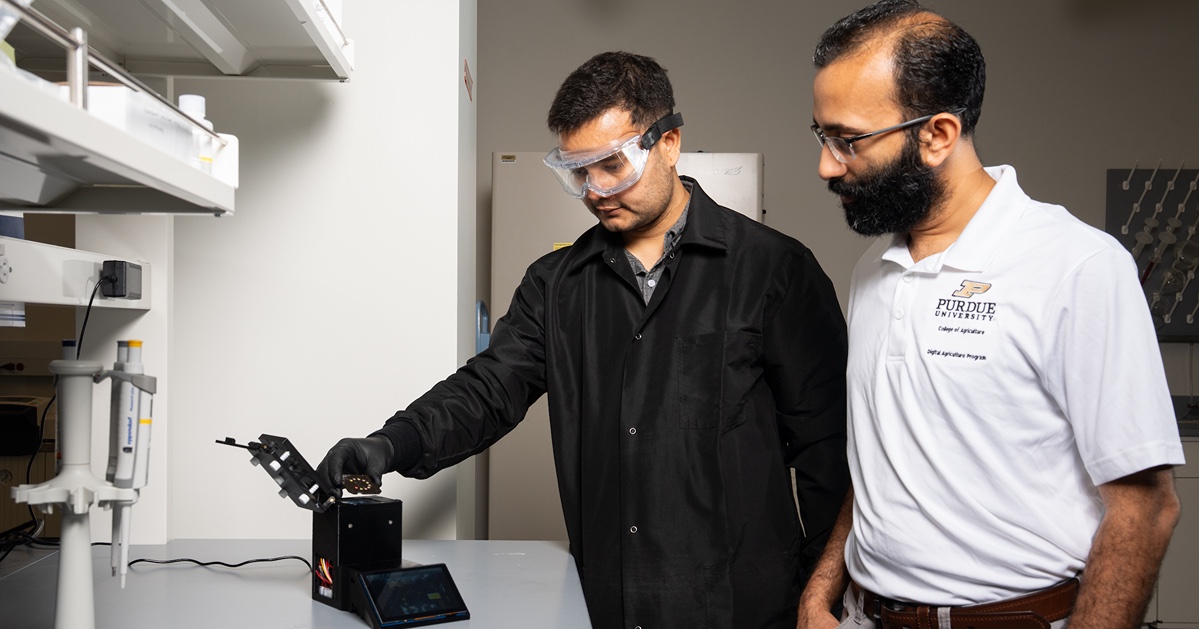 Two men, one wearing a lab coat, examine a scientific device in a laboratory setting.