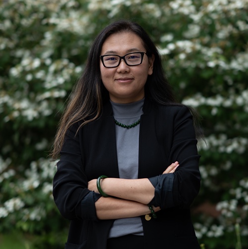 A woman wearing glasses and a black jacket stands in front of a large tree, smiling at the camera.