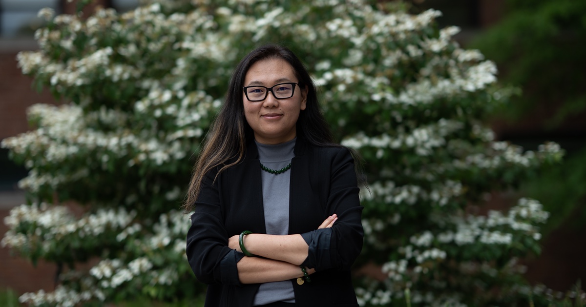 A woman wearing glasses and a black jacket stands in front of a large tree, smiling at the camera.