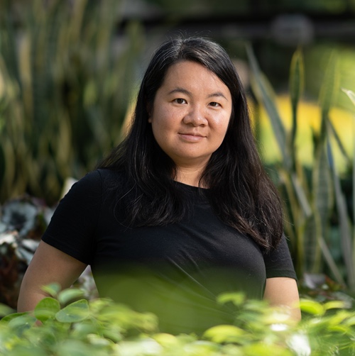 A woman stands in a greenhouse, surrounded by greenery and natural light.