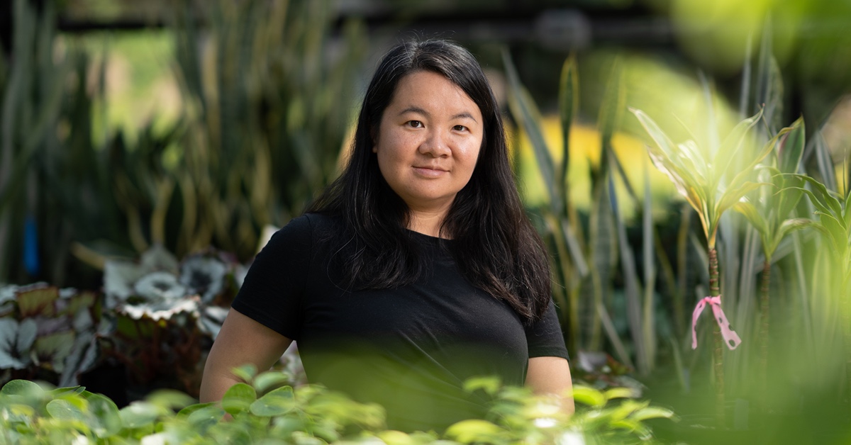 A woman stands in a greenhouse, surrounded by greenery and natural light.