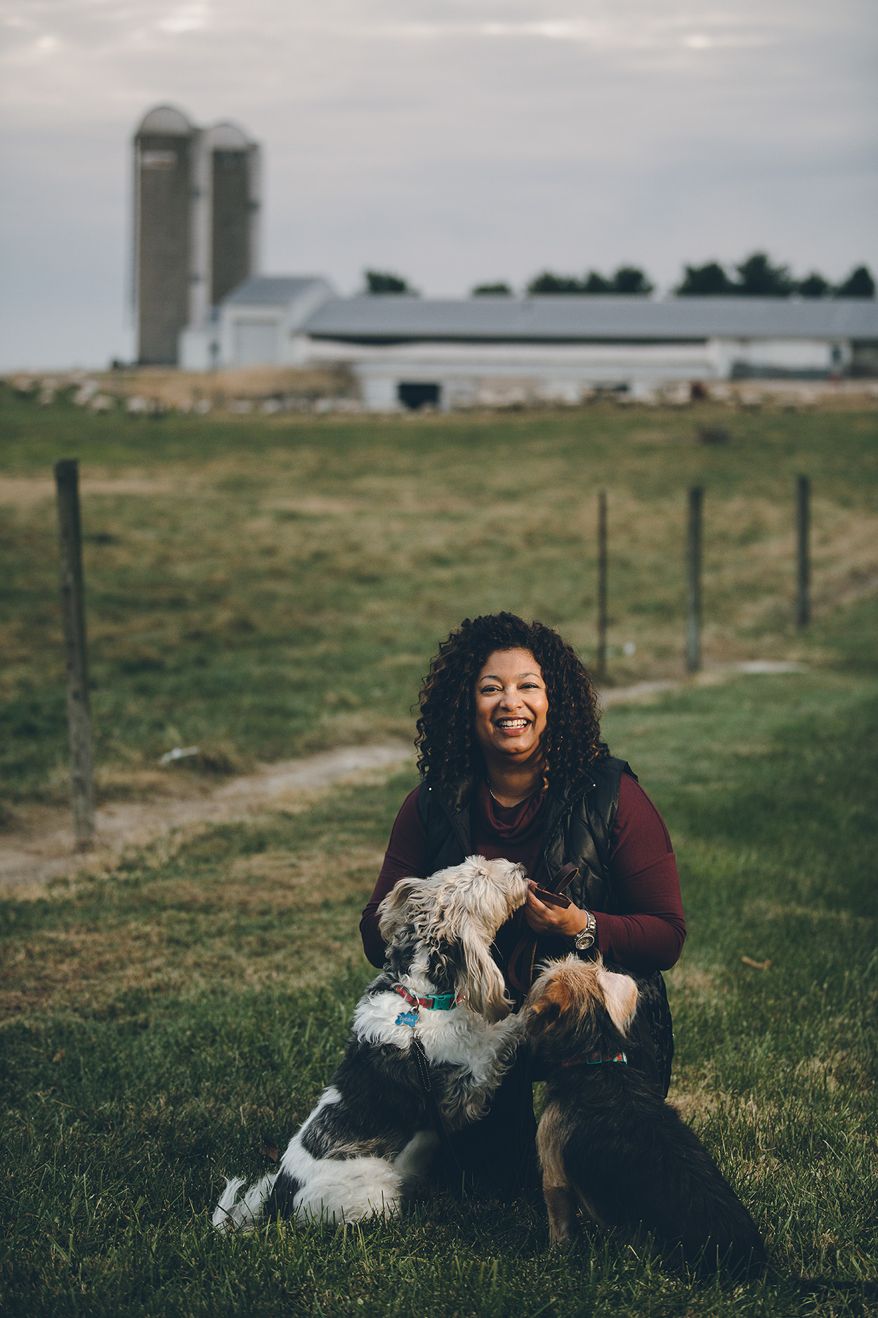 Candace Croney kneels in a grassy field with two dogs, smiling toward the camera. A livestock barn and silos are visible in the background.