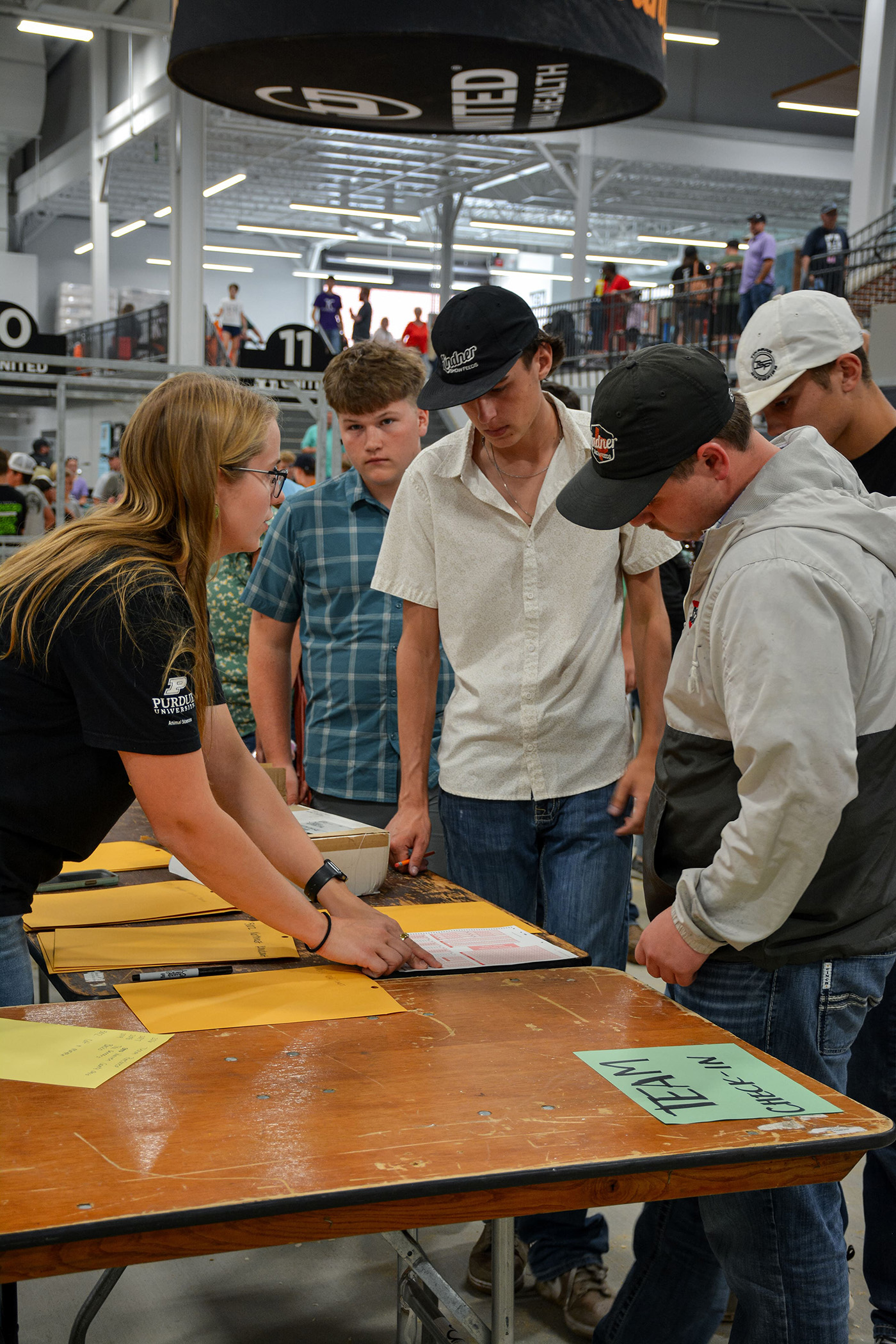 Group of people gathered around a table labeled 'TEAM CHECK-IN' with yellow envelopes and papers, including a person in a Purdue University shirt pointing at a document.