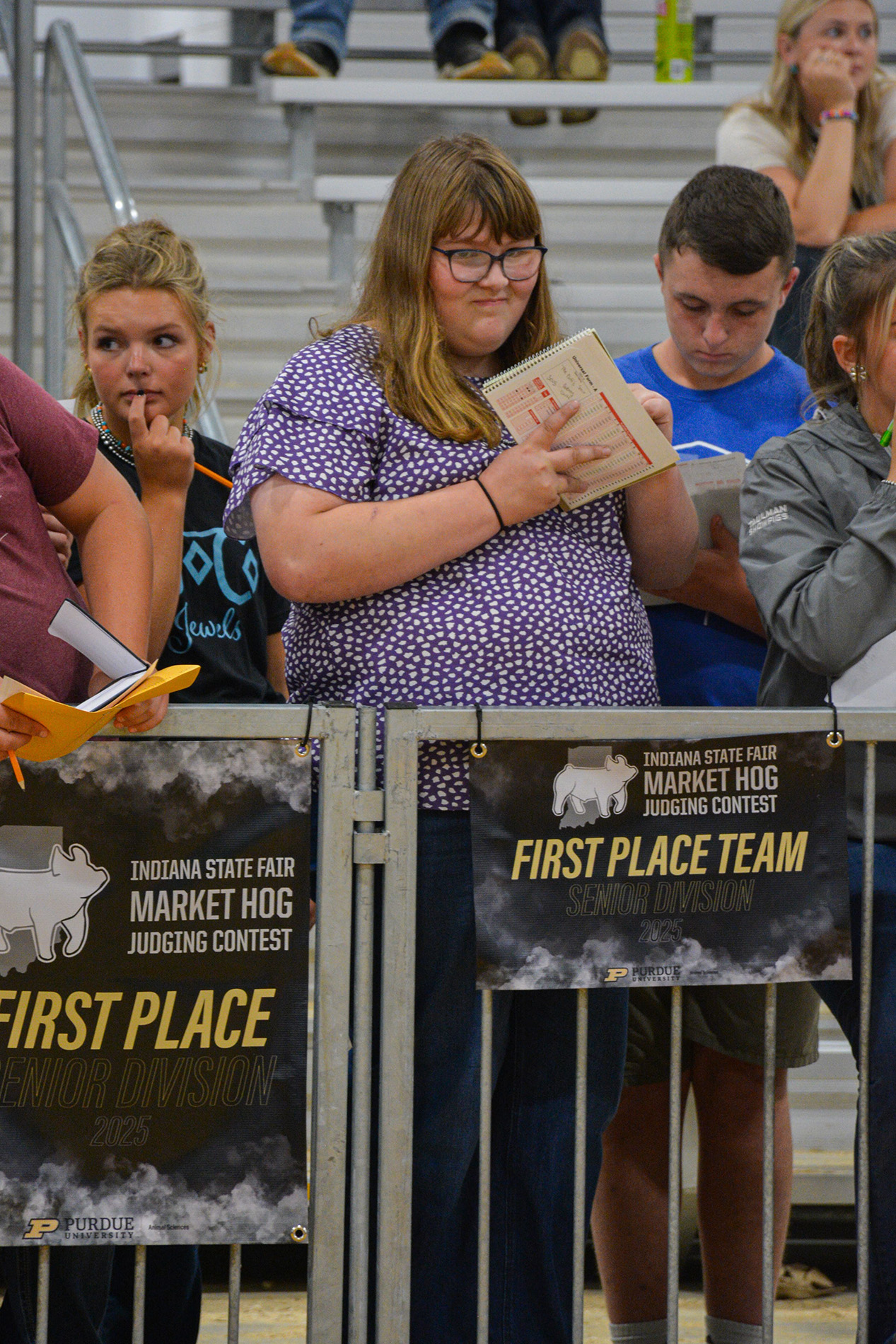 Group of people standing behind a metal barrier displaying two banners for the Indiana State Fair Market Hog Judging Contest.