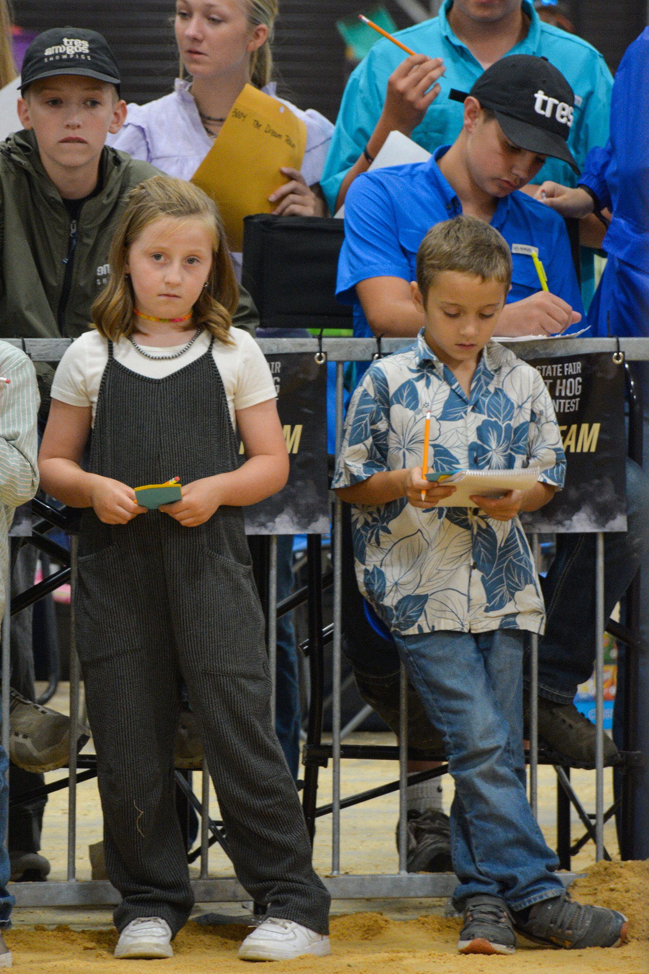 Two children holding notebooks and pencils in front of a metal barrier at the State Fair Hog Contest, with older kids and event signage in the background.